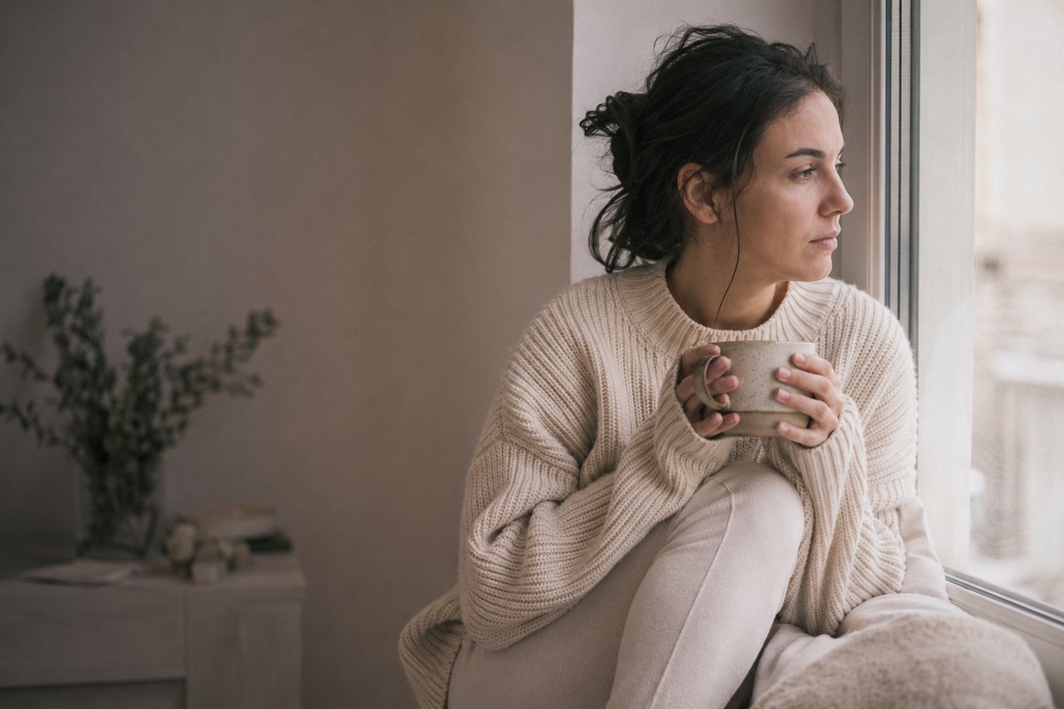 A woman in a cream sweater sits by a window in early morning light, holding a mug, looking out